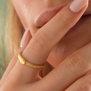 May include: A gold snake ring with a textured surface. The ring is worn on a finger.