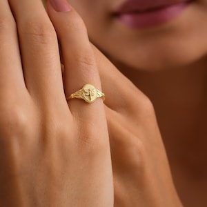 May include: A gold-colored ring with an oval-shaped face featuring a dragonfly design. The ring has a delicate band and is worn on a finger. The dragonfly is centered on the oval, surrounded by a beaded border.