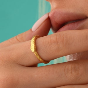 May include: A gold snake ring with a textured finish. The ring is worn on a finger.