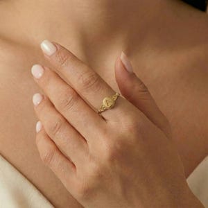 Woman wearing a gold initial signet ring with the letter A on an oval face, hand resting near her chest.
