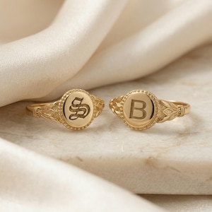 May include: Two gold signet rings with engraved initials. One ring features the letter "S" in an ornate font, and the other displays the letter "B". The rings have a classic design with a beaded border and decorative shoulders.