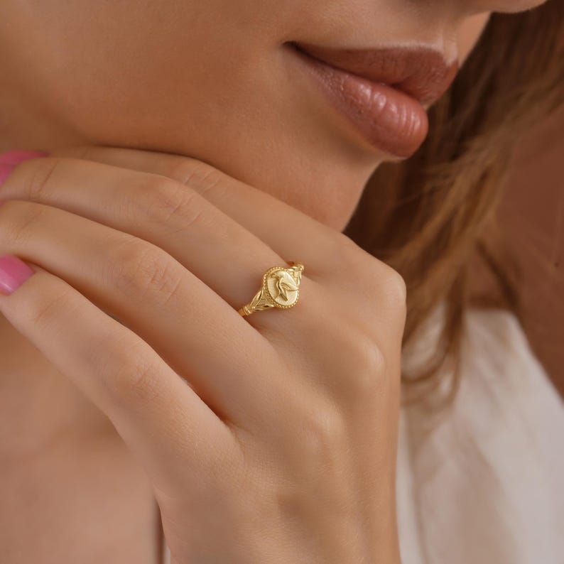 K&ouml;nnte beinhalten: Ein goldener Ring mit einem detaillierten Design, das ein zentrales ovales Emblem aufweist. Der Ring hat ein zartes, verziertes Band und eine strukturierte Oberfl&auml;che. Der Ring wird an einem Finger getragen.