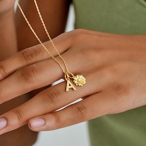 May include: A gold-toned necklace featuring a delicate chain with two charms: a capital letter A pendant and a small, textured flower charm with a tiny, clear, central stone. The metal has a polished finish, and the charms hang side by side.