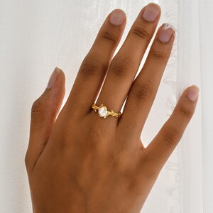 May include: A gold-colored ring featuring a central, oval-shaped, white gemstone. The ring's band is designed with a delicate, nature-inspired motif, resembling intertwined branches and leaves. The ring is displayed on a hand.