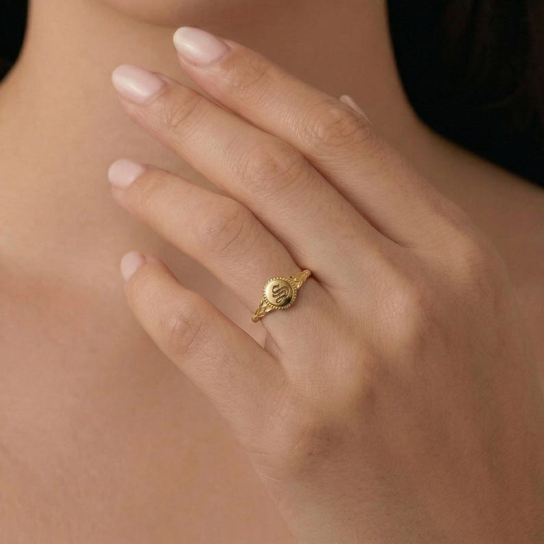 Model's hand wearing a gold initial signet ring with an embossed letter, posed against her chest.