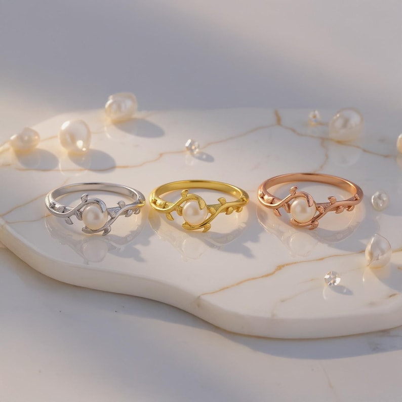 May include: Three rings with a pearl center and a delicate vine design. The rings are silver, gold, and rose gold. Scattered pearls and small crystals surround the rings on a white marble surface.