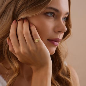 May include: A gold-colored ring with a circular design is worn on a finger. The ring is the focal point, with the person's face and hand in the background. The ring's design is visible, and the metal has a shiny appearance.