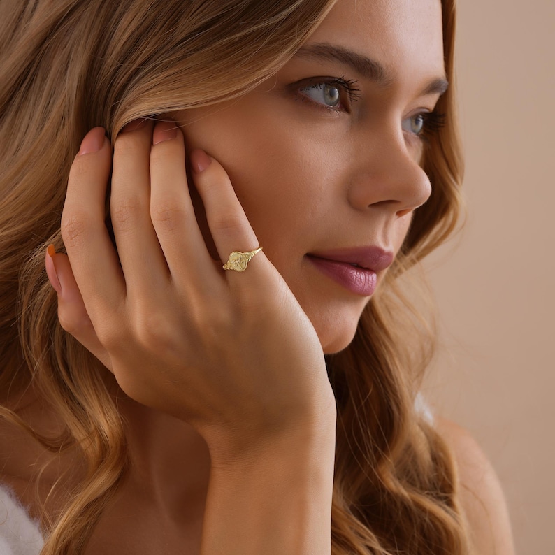 May include: A gold-colored ring with a circular design is worn on a finger. The ring is the focal point, with the person's face and hand in the background. The ring's design is visible, and the metal has a shiny appearance.