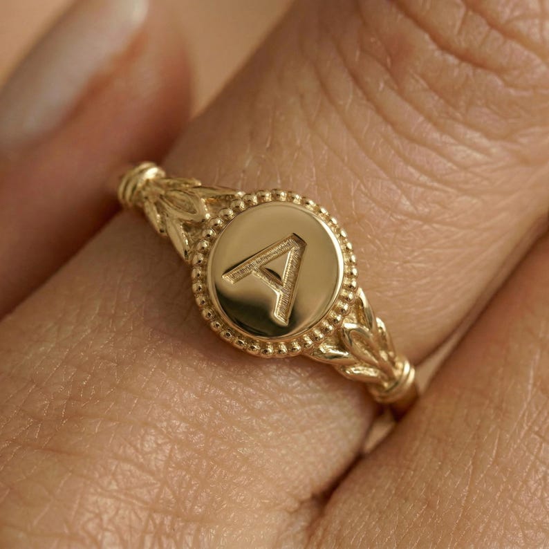 Close-up of a gold initial signet ring with the letter A on a circular face and beaded border, worn on a finger.