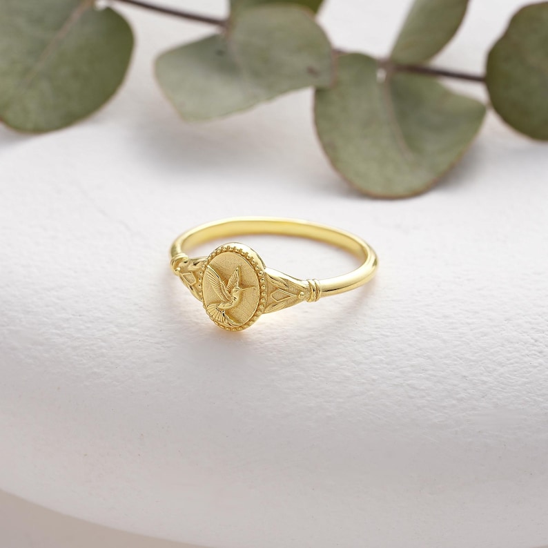 A gold ring with a detailed oval-shaped design featuring a small hummingbird in flight. The ring is simple and elegant.