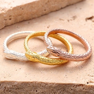 May include: Three snake-shaped rings in silver, gold, and rose gold. The rings are stacked on top of each other.