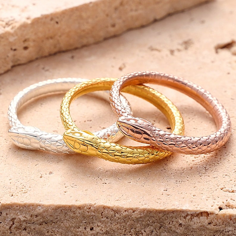 May include: Three snake-shaped rings in silver, gold, and rose gold. The rings are stacked on top of each other.