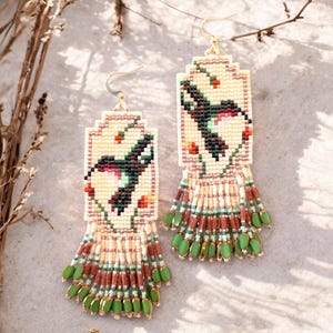 Può includere: Un paio di orecchini di perline con un design rettangolare, con un motivo a uccello in nero, rosso e verde. Gli orecchini hanno uno sfondo color crema e una frangia di perline verdi e marroni. I ganci dorati completano il design.