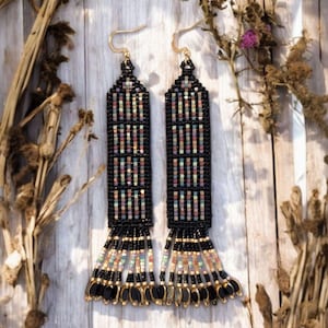 May include: A pair of long, beaded earrings with a rectangular design. The earrings are mainly black with colourful accents and gold-coloured hooks and fringe. The earrings are displayed on a light wooden surface.