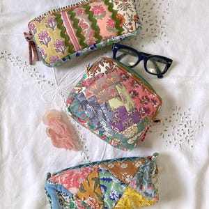 May include: Three rectangular and square zipped pouches made from assorted floral patterned fabric scraps. The top pouch features green rickrack trim, the middle pouch has a patchwork design, and the bottom pouch displays a triangular quilt pattern.