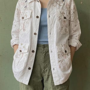 May include: A white button-down shirt featuring intricate floral embroidery in soft pink and brown tones, a classic collar, long sleeves, and four front pockets with button closures. The garment is worn over a light blue tank top and olive green trousers.