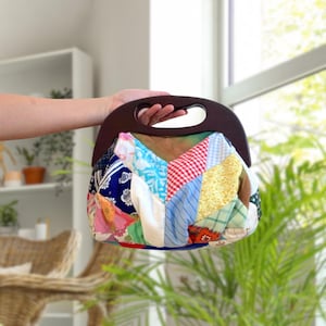 May include: A patchwork handbag with a dark brown wooden handle. The bag is made of various fabric scraps in a variety of colors and patterns, including red gingham, blue floral, and yellow. The bag is held up by a person's hand.