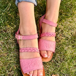 May include: Pink leather sandals with braided straps and floral embossed detailing. The sandals have a brown leather sole and an adjustable ankle strap with a gold buckle.