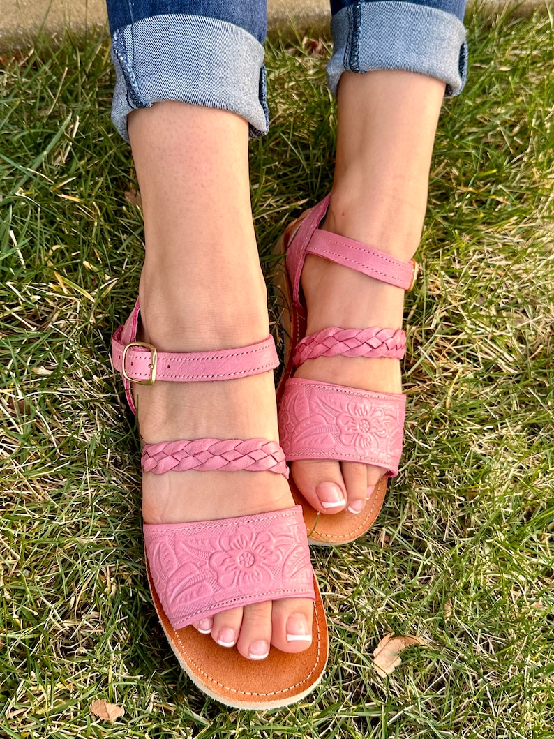 May include: Pink leather sandals with braided straps and floral embossed detailing. The sandals have a brown leather sole and an adjustable ankle strap with a gold buckle.