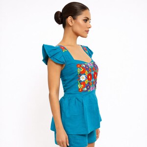 May include: A turquoise two-piece outfit featuring a top and shorts. The top has a square neckline, ruffled sleeves, and is adorned with colorful floral embroidery. The shorts are a matching turquoise color. The outfit is made of a textured fabric.