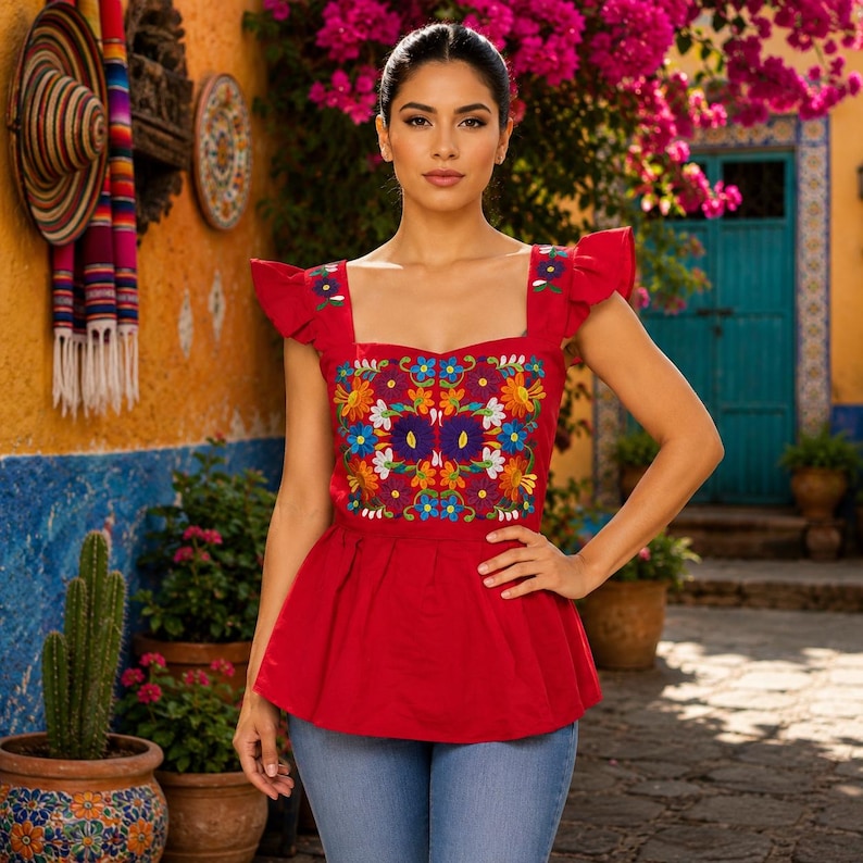 May include: A red embroidered top with a square neckline and ruffled sleeves. The top features a vibrant floral design in various colors. The model is wearing blue jeans. The background includes a colorful wall and plants.
