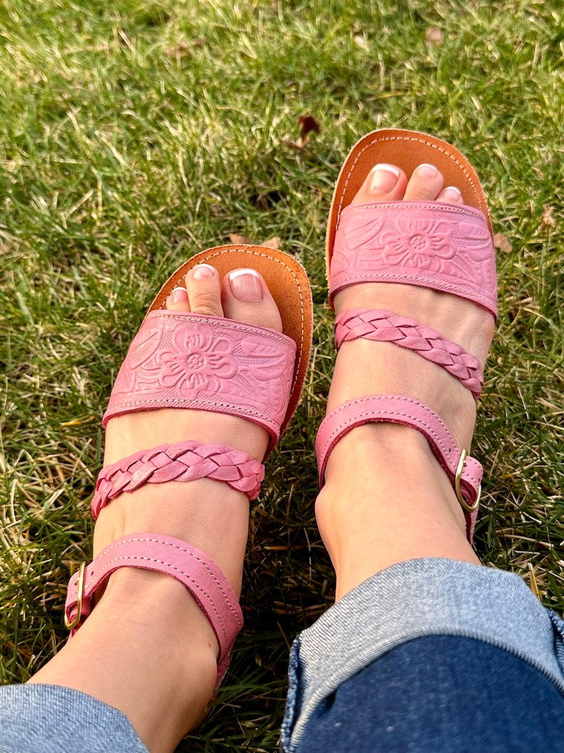 May include: A pair of pink leather sandals with braided straps and floral detailing. The sandals have an ankle strap with a buckle closure.