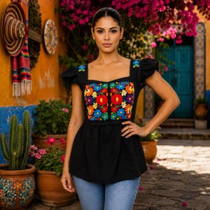 May include: A black top with a square neckline and short sleeves. The top features vibrant, colorful floral embroidery on the bodice. The model is wearing blue jeans. The background includes a cactus, flowers, and a colorful wall.