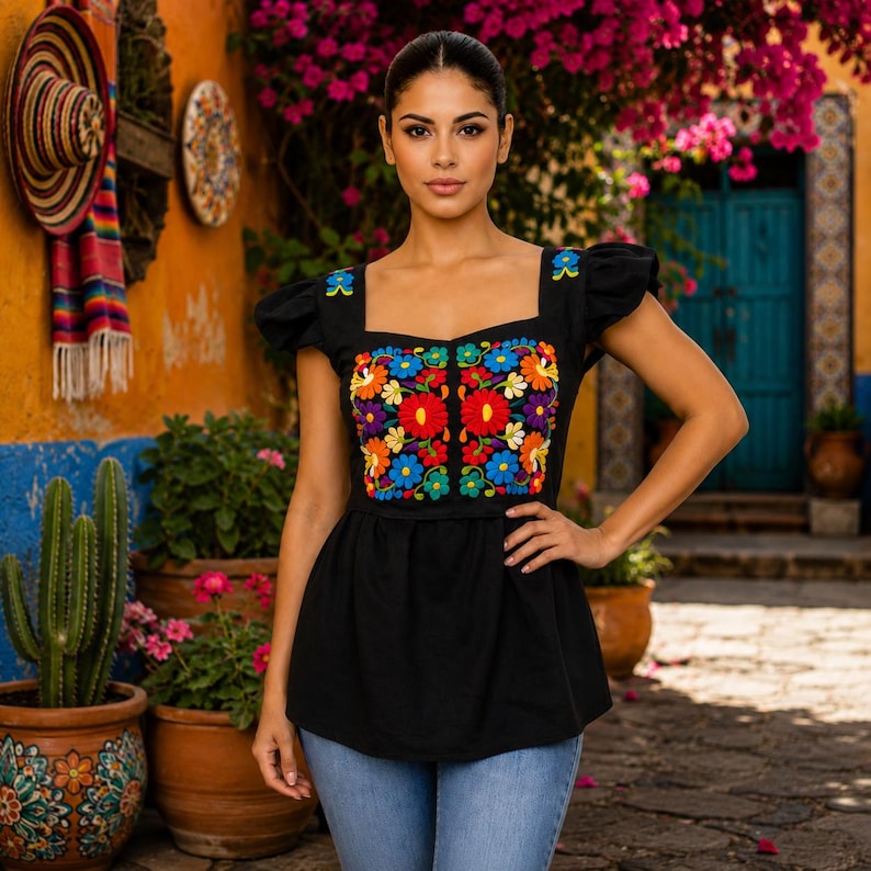May include: A black top with a square neckline and short sleeves. The top features vibrant, colorful floral embroidery on the bodice. The model is wearing blue jeans. The background includes a cactus, flowers, and a colorful wall.