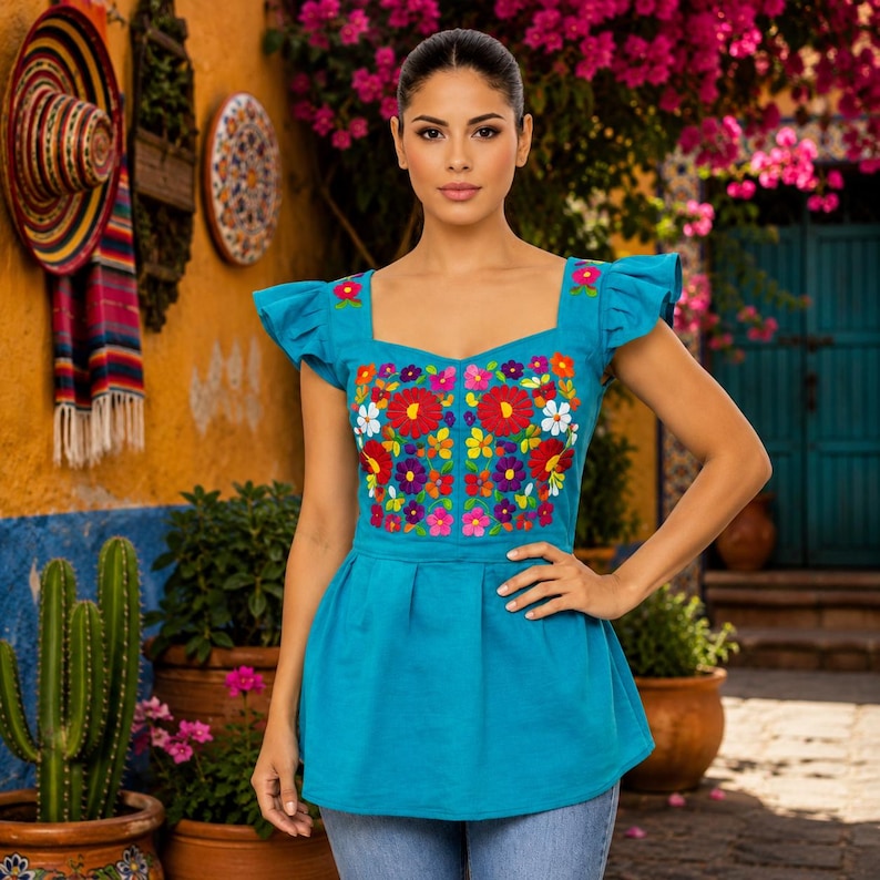 May include: A turquoise embroidered top with a square neckline and ruffled sleeves. The top features a vibrant floral design in red, white, yellow, purple, and blue. The top is paired with blue jeans.