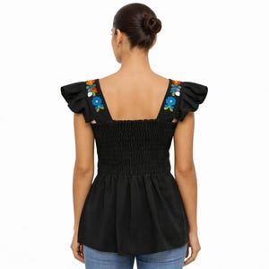 May include: Black top with floral embroidery on the shoulder straps. The top features a square neckline, ruffled sleeves, and a smocked bodice. The floral embroidery includes blue, orange, and white flowers. The bottom of the top has a flared design.