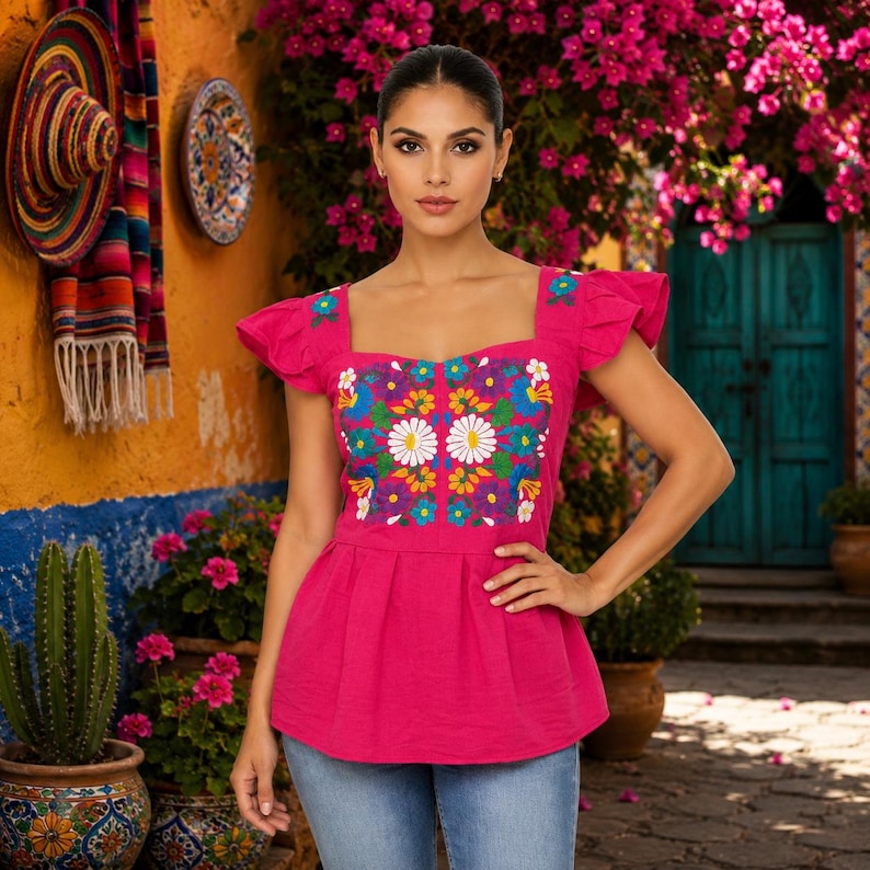 May include: A vibrant pink top with a square neckline and ruffled sleeves. The top features colorful floral embroidery on the bodice. The model is wearing blue jeans. The background includes a colorful wall, flowers, and a door.