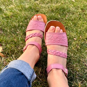 May include: A pair of pink leather sandals with braided straps and floral embossed designs. The sandals have a closed toe and an ankle strap with a buckle.