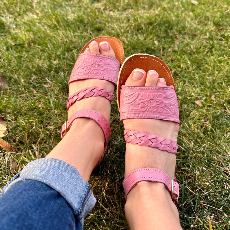 May include: A pair of pink leather sandals with braided straps and floral embossed designs. The sandals have a closed toe and an ankle strap with a buckle.