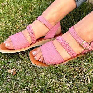May include: A pair of pink leather sandals with braided straps and a buckle closure. The sandals are on a grassy surface.