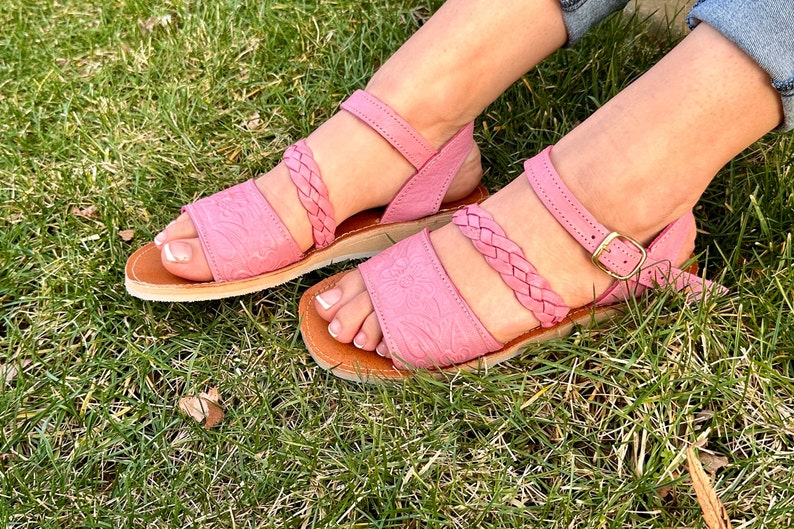 May include: A pair of pink leather sandals with braided straps and a buckle closure. The sandals are on a grassy surface.