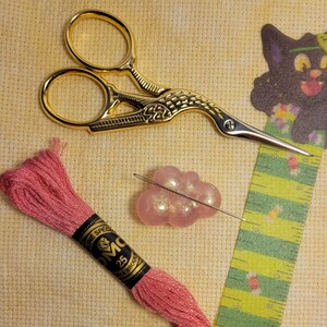 May include: A pink embroidery floss skein, a pair of gold-coloured scissors with a bird design, a pink and white beaded cloud charm, and a green and yellow ruler with a black cat design.