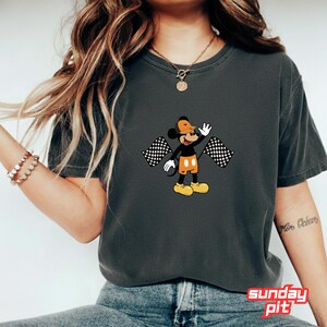 May include: Dark gray t-shirt featuring a cartoon Mickey Mouse character waving and holding two checkered flags. Mickey is wearing an orange hat, shorts, and yellow shoes. The text "sunday pit" is at the bottom.