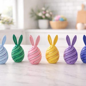 May include: Six colorful Easter egg decorations with bunny ears. The eggs are in shades of blue, green, pink, yellow, purple, and dark blue. Each egg has a spiral design. The decorations are on a white surface.