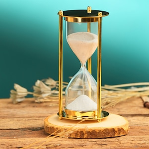 May include: An hourglass with a gold-coloured frame and clear glass bulbs filled with white sand. The hourglass sits on a wooden base, with a teal background. The hourglass is approximately 15 cm tall.
