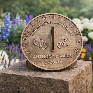 May include: Bronze sundial marking a 10th wedding anniversary. It displays Roman numerals, the names "Anthony & Karis," and the date 7-20-14. Two heart designs are included. The sundial is set on a stone base, with a garden in the background.