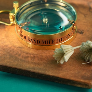 May include: A vintage-style brass compass with a clear glass top and the inscription "THOUSAND MILE JOURNEY." The compass features a gold-colored ring and a small loop. A dried flower rests beside the compass on a brown surface.