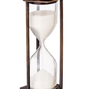 Puede incluir: Un reloj de arena de estilo antiguo con un marco de metal color bronce y bombillas de vidrio transparente. El reloj de arena contiene arena blanca. El marco tiene una base y una parte superior cuadradas, con soportes verticales. El reloj de arena mide unos 15 cm de alto.