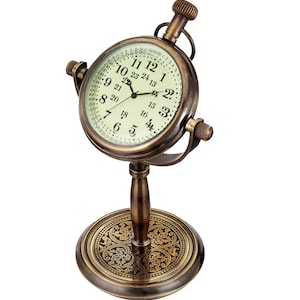 May include: A brass desk clock with a round face and a stand. The clock face has a green background with black numbers and hands. The stand has a decorative base with a floral pattern.