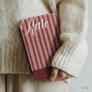 May include: A rectangular tablet case featuring a vertical striped pattern in alternating shades of terracotta and light pink. The name Kate is printed in a white cursive script across the upper portion. A solid dark maroon band borders the bottom.
