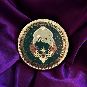 Peut inclure: Épingle ronde à bordure dorée et centre vert foncé. Le motif comprend un paysage de montagne, des bois de cerf et une étoile, avec des détails rouges et blancs. L'épingle est posée sur un tissu de satin violet.