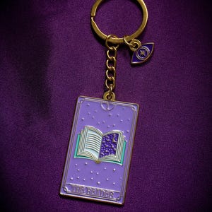 May include: A gold-coloured keyring with a purple tarot card pendant. The pendant features an open book design with a starry night scene and the text "THE READER". An additional charm with an eye design is attached.