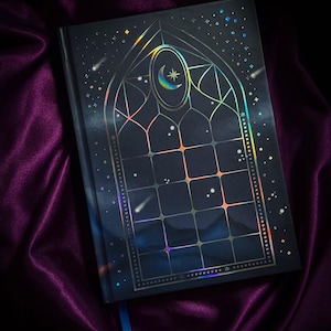 Può includere: Un diario blu scuro con un design celeste. La copertina presenta un design a vetrata olografica con una luna crescente e stelle. Il diario è appoggiato su un tessuto di raso viola.