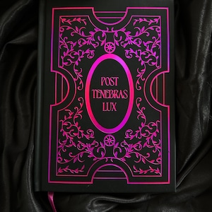 Op de afbeelding: Zwart notitieboek met een roze en paars bloemmotief en de tekst "POST TENEBRAS LUX". De kaft heeft een sierlijk, reliëfpatroon. Een roze lint-bladwijzer is zichtbaar.
