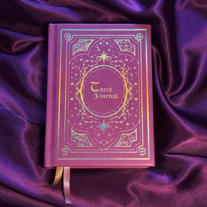 Può includere: Un diario magenta con le parole "Tarot Journal" in lettere dorate. La copertina presenta un design ornato con stelle ed elementi botanici. Un segnalibro a nastro dorato e rosa è visibile.