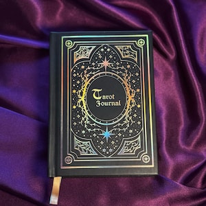 May include: Black Tarot Journal with gold foil accents. The cover features the words "Tarot Journal" in a decorative font, surrounded by celestial designs. A gold ribbon bookmark is visible. The journal is set against a purple satin background.